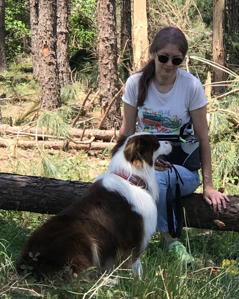 Leigh sitting on a pine trunk felled by the April 2020 tornado, in the East Texas piney woods, with a dog beside her and storm-broken pines behind